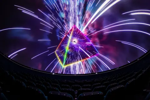 A projection of a glowing pyramid and light streaks inside a domed theater with rows of seats.