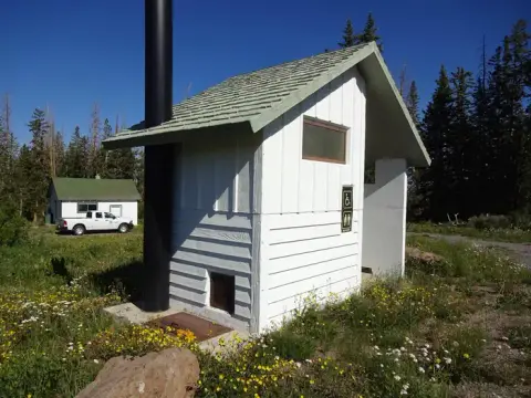 A small white outdoor restroom building stands in a grassy area with trees and a clear blue sky.