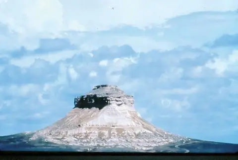 A large isolated rock formation stands beneath a partly cloudy sky.