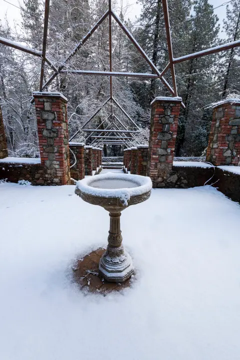 A snow-covered birdbath sits in an outdoor structure with brick pillars and a metal frame surrounded by trees.
