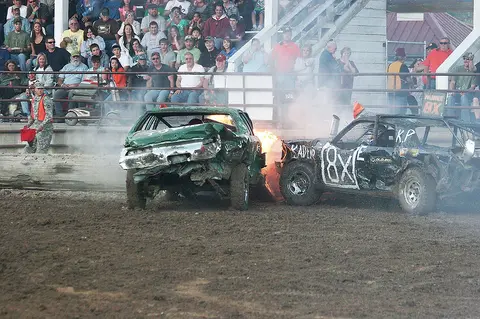 Two damaged cars are crashing in a demolition derby in front of a crowd of spectators.