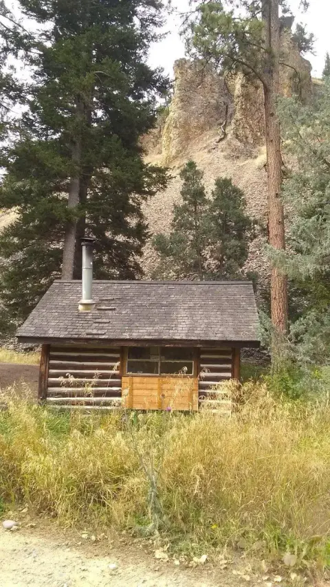 A small log cabin stands among trees and tall grass with a rocky hill in the background.