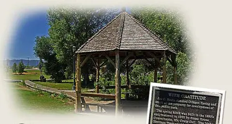 A wooden gazebo stands in a park with a commemorative plaque in the foreground.