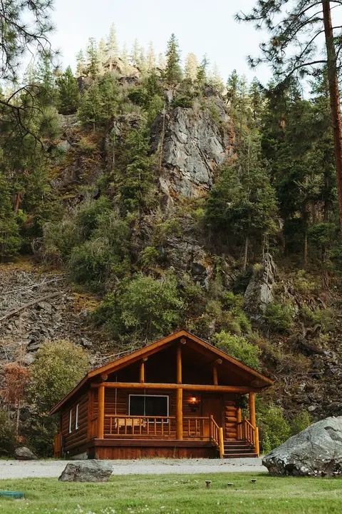 A wooden cabin sits at the base of a steep, rocky, tree-covered hill.