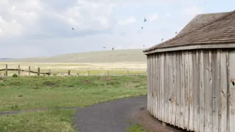 A wooden building stands beside a path overlooking a grassy field and distant hills.