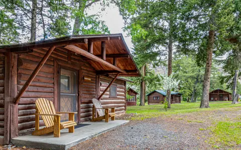 A rustic wooden cabin with two chairs on the front porch stands surrounded by trees.