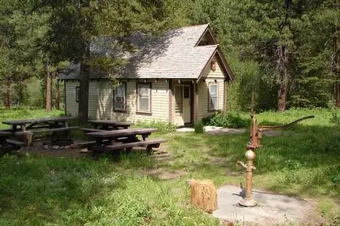 A small cabin stands surrounded by trees with picnic tables and a water pump in the foreground.