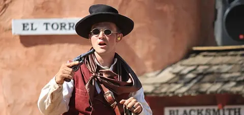 A man dressed in cowboy attire holds a revolver outdoors.