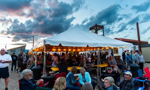 People gather under a large white tent at an outdoor gathering during sunset.