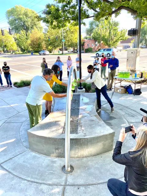 People gather around an outdoor water feature to fill bottles while others take photos.