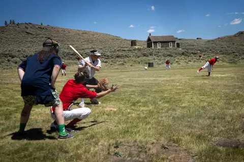People play vintage baseball on a grassy field in front of a rustic building.