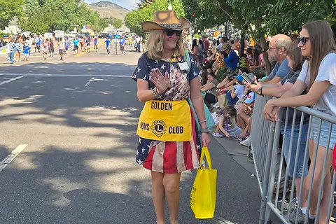 A woman in patriotic clothing waves to a crowd gathered along a street for a parade.