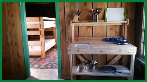 A rustic wooden kitchen counter stands in an interior room with a bunk bed visible through a doorway.