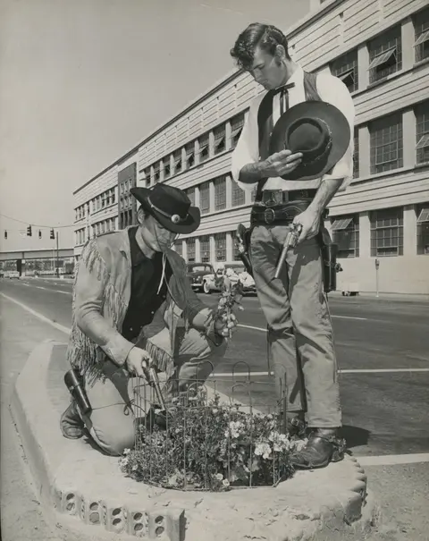 Black and white photo of two cowboys in front of a modern building looking at flowers in a street planter.