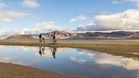 Three people walk along a lakeshore with mountains in the background.