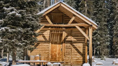 A wooden cabin sits surrounded by snow and evergreen trees.