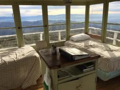 An interior view of a room with large windows overlooking a mountainous landscape, two beds, and a central table.