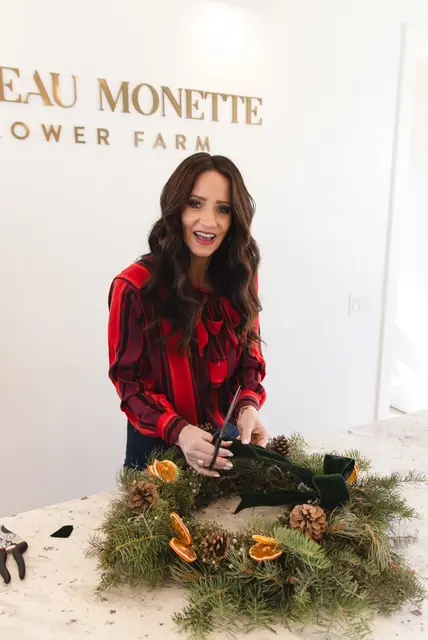 A woman smiling while making a holiday wreath inside a room.