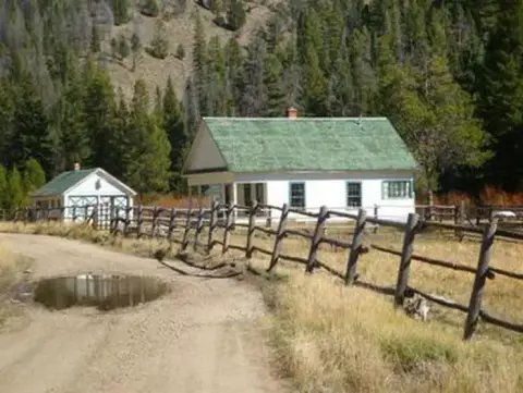 A dirt road leads past a white farmhouse and barn with green roofs nestled against a forested hillside.