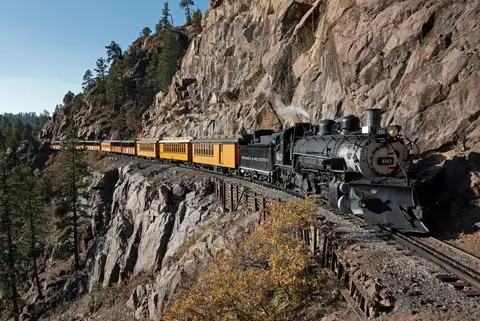 A steam train travels along a rocky cliffside track.