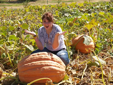 A young woman poses excitedly in a pumpkin patch with large pumpkins.