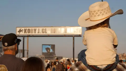 A child wearing a cowboy hat sits on someone's shoulders at an outdoor arena event.