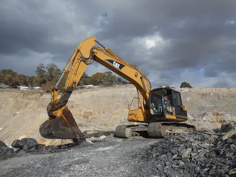 A yellow excavator working at a rocky quarry under a cloudy sky.