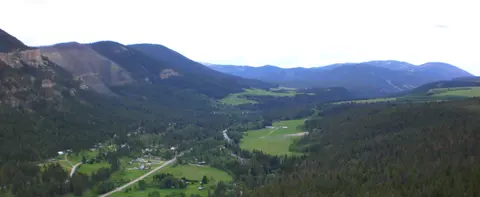 A wide landscape view of a green valley surrounded by forested mountains under an overcast sky.