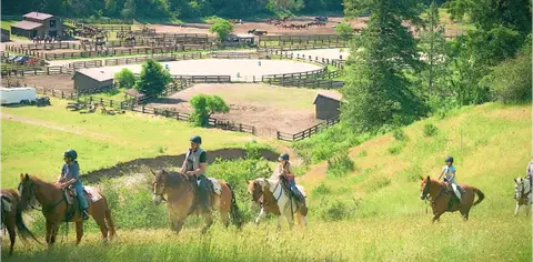 People ride horses on a grassy hill with a ranch facility in the background.