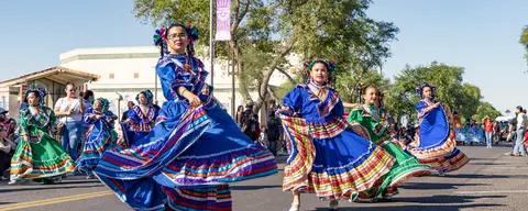 Performers in colorful traditional dresses dance in a street parade.