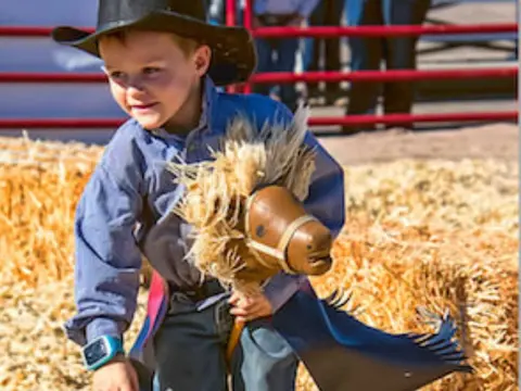 A young boy dressed as a cowboy plays with a hobby horse near hay bales.