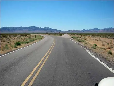 A paved road curves through a desert landscape with distant mountains under a blue sky.
