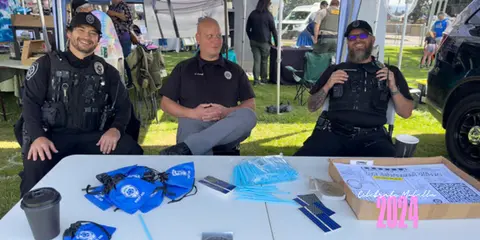 Three officers sit at a table outdoors at a community event.