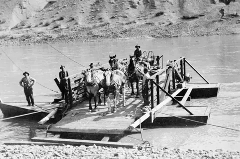 A historic black and white photograph showing a cable ferry carrying horses and people across a river.