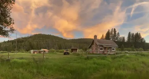 A scenic view of a log cabin in a grassy field with forested hills under a cloudy sunset sky.