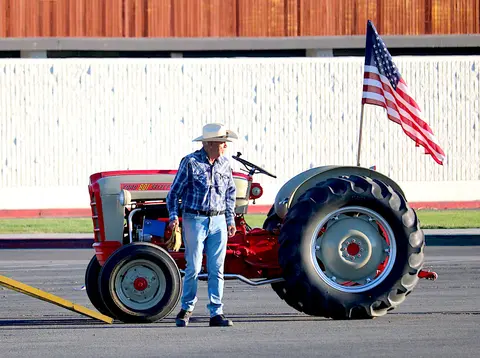 A man wearing a cowboy hat stands next to a red tractor with an American flag attached to it.