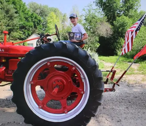 A young person sits on a red tractor holding an award, with American flags attached to the back.