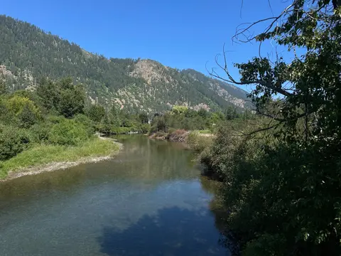 A river flows through a lush green valley with forested mountains under a clear blue sky.