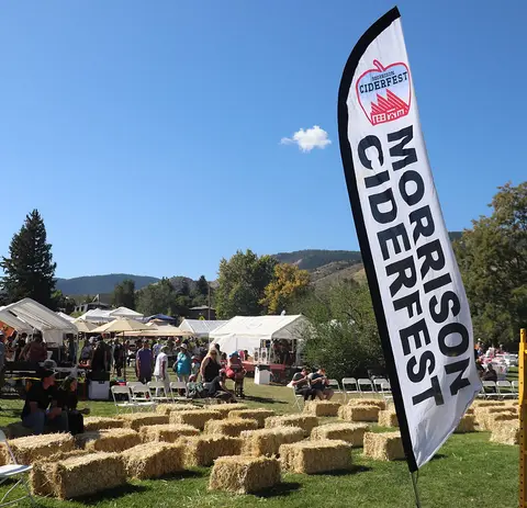 An outdoor festival features a large banner, hay bales, and white tents on a grassy field.