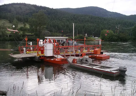 A small orange ferry boat sits on a river with mountains in the background.