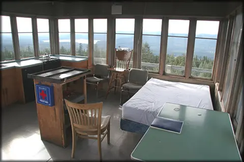 An interior view of a fire lookout cabin with a bed, table, and windows showing a scenic view.