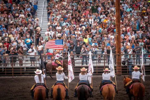 Several horse riders carrying flags stand in an arena before a large crowd of spectators.