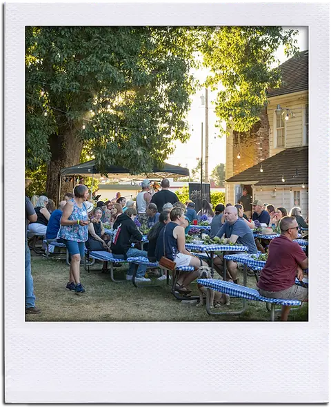A crowd of people seated at picnic tables outdoors near a building and trees.