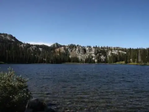 A scenic view of a calm lake surrounded by trees and mountains under a blue sky.