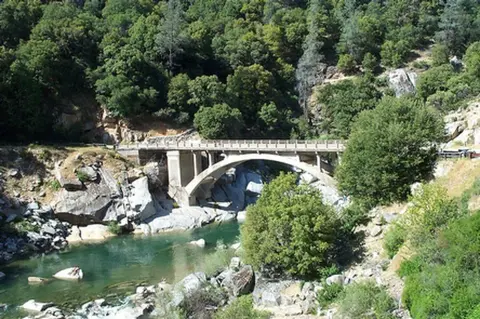 A concrete arch bridge spans a river surrounded by trees and rocky terrain.