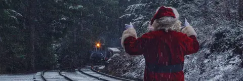Santa Claus stands next to snowy train tracks as a train approaches in the forest.