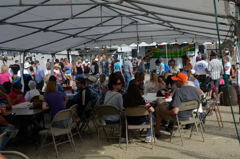 People sit at tables under a large white tent at an outdoor concert event.