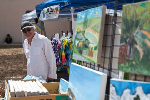 A smiling artist stands at an outdoor art market booth displaying paintings and tie-dye items.