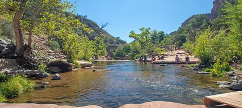 People relax along a shallow stream surrounded by trees and rocky banks.
