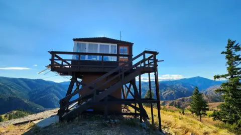 A wooden fire lookout tower stands on a mountain peak with distant ranges in the background.